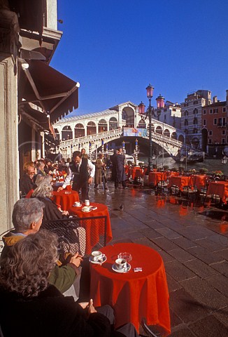 Tourists enjoy coffee at waterside caf with view to  Rialto bridge on a sunny autumnal day Venice Italy