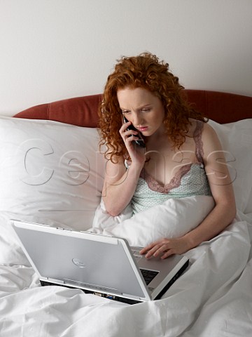 Young woman in bed with laptop computer and talking on her mobile phone