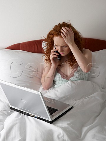 Young woman in bed with laptop computer and talking on her mobile phone