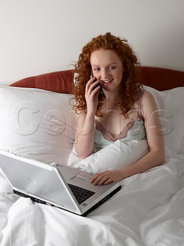 Young woman in bed with laptop computer and talking on her mobile phone