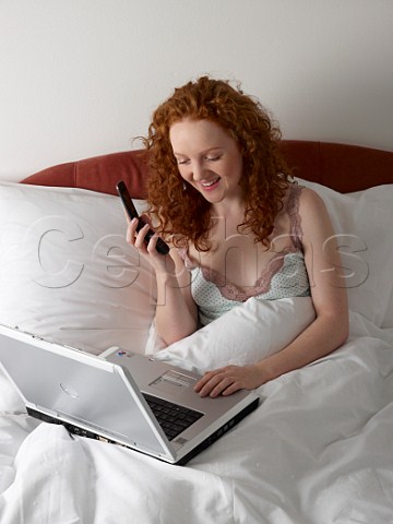 Young woman in bed with laptop computer and talking on her mobile phone