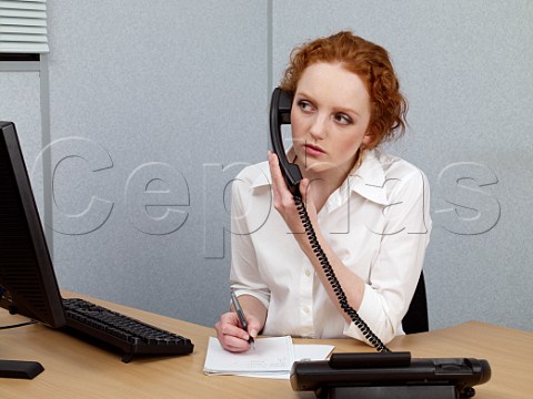 Young woman at her office desk making notes whilst on telephone