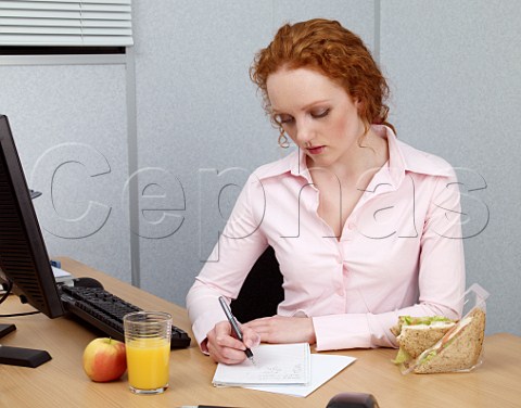 Young woman at her office desk making notes during her lunch break Chicken salad sandwich glass of orange juice apple