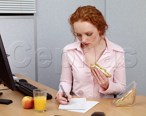 Young woman at her office desk making notes during her lunch break Chicken salad sandwich glass of orange juice apple