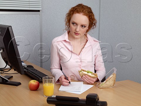 Young woman at her office desk making notes during her lunch break Chicken salad sandwich glass of orange juice apple