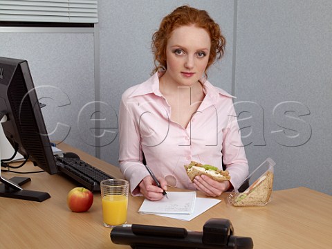 Young woman at her office desk making notes during her lunch break Chicken salad sandwich glass of orange juice apple