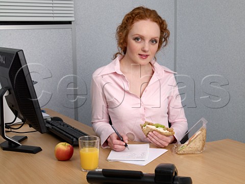 Young woman at her office desk making notes during her lunch break Chicken salad sandwich glass of orange juice apple