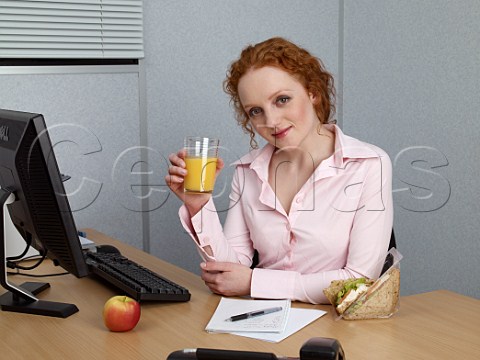 Young woman at her office desk making notes during her lunch break Chicken salad sandwich glass of orange juice apple