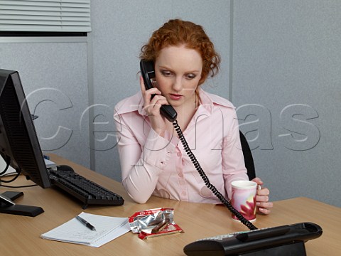 Young woman on telephone at her office desk with mug of tea and a KitKat