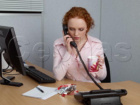 Young woman on telephone at her office desk with mug of tea and a KitKat