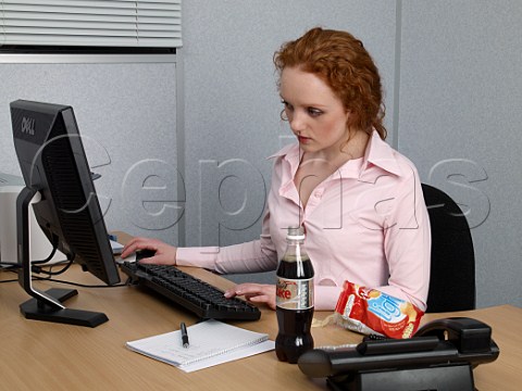Young woman working on computer at her office desk bottle of diet coke and bag of lofat crisps