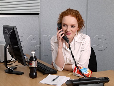 Young woman on telephone at her office desk with bottle of diet coke and bag of lofat crisps