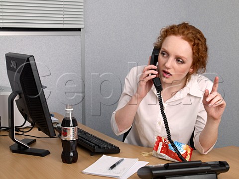 Young woman on telephone at her office desk with bottle of diet coke and bag of lofat crisps trying to attract the attention of a colleague