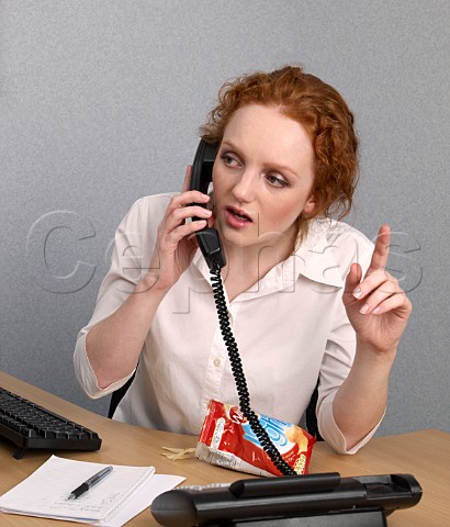 Young woman on telephone at her office desk with bag of lofat crisps trying to attract the attention of a colleague