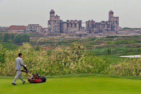 Worker mowing the lawn at Junding winery golf course with old cement factory in distance near Penglai Shandong Province China